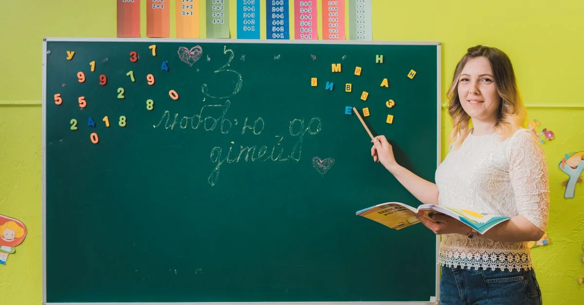 Une enseignante pointant un tableau noir avec des chiffres dans une salle de classe colorée.